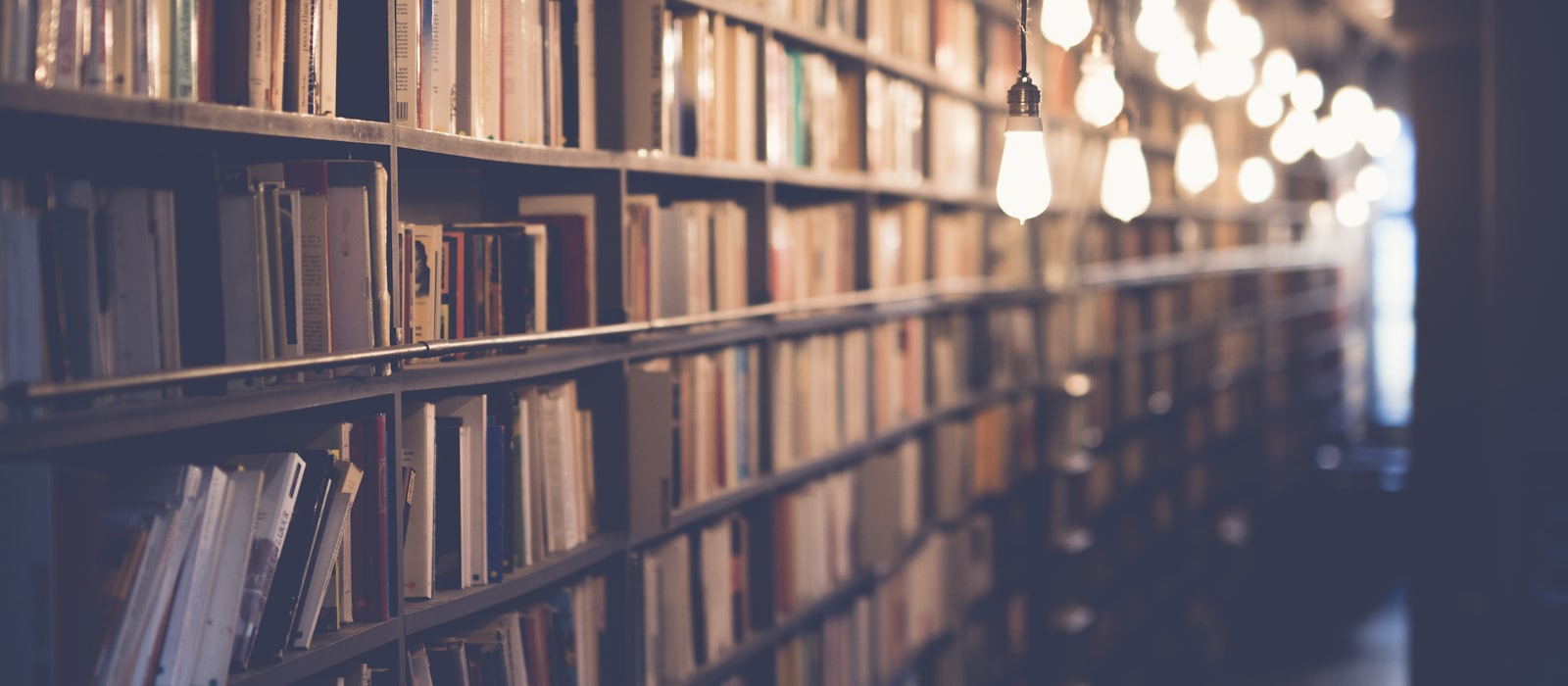 A long library aisle of packed bookshelves lit by hanging bulbs
