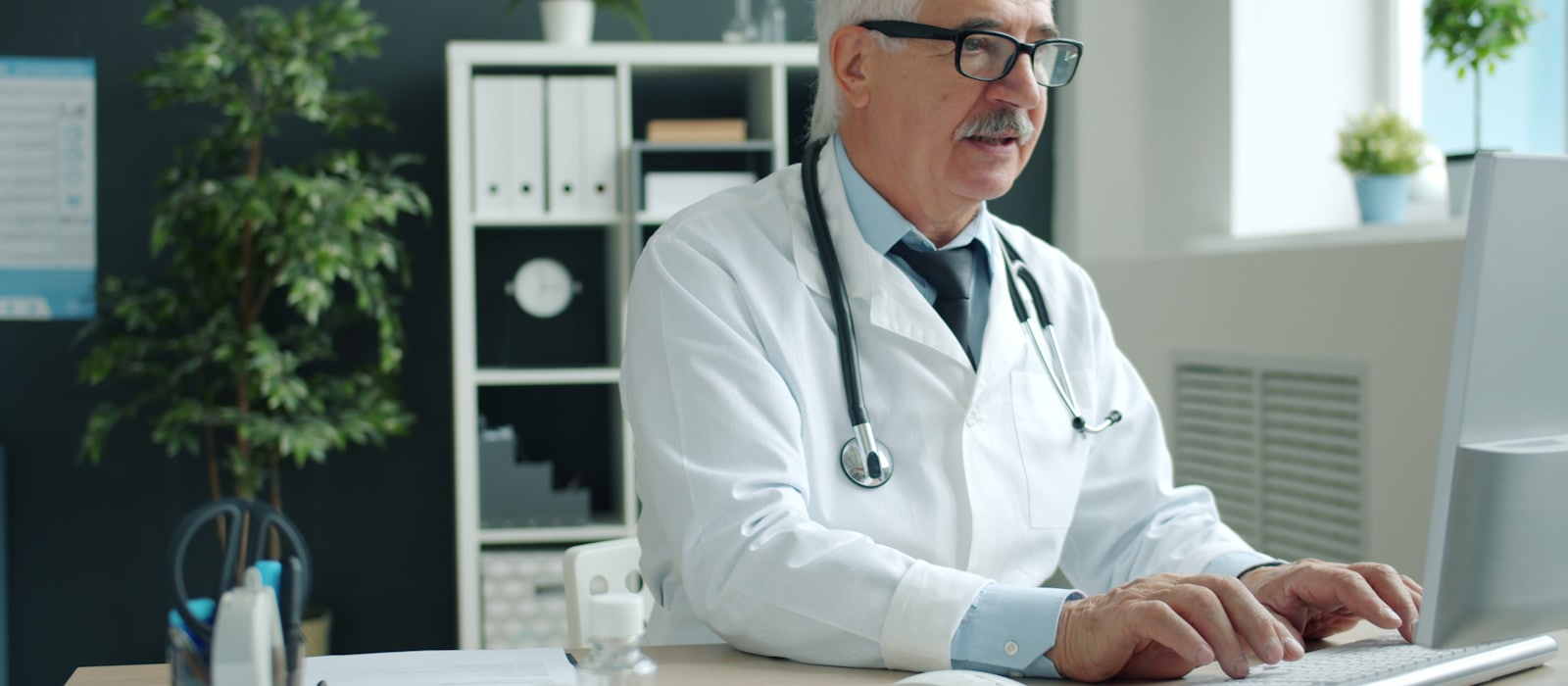 A doctor in a white coat with a stethoscope working at a computer in a consulting room