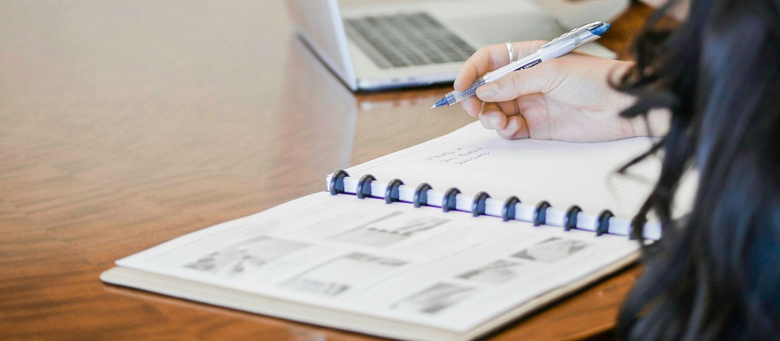 A person writing in a spiral-bound workbook at a desk beside a laptop
