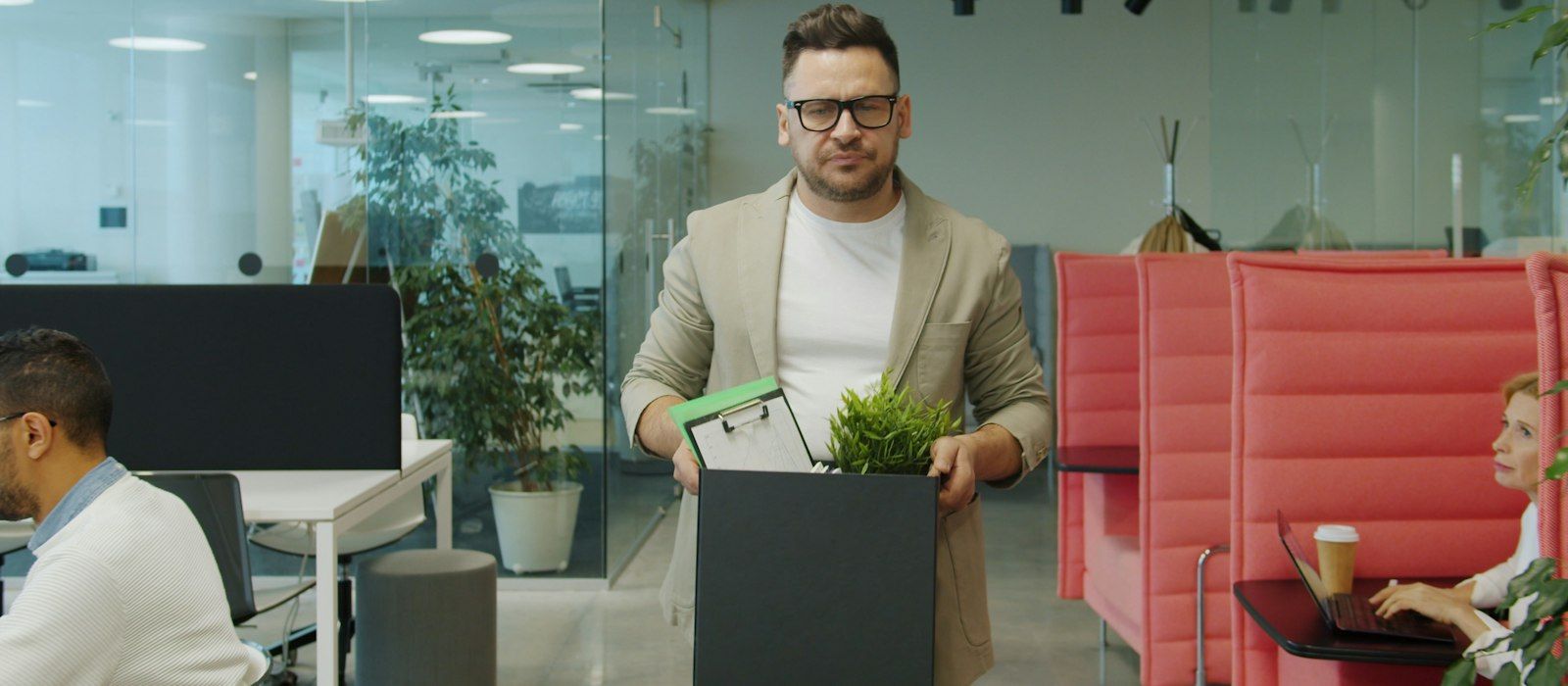 A man in glasses walks through an open-plan office carrying a box of his belongings while colleagues work at their desks