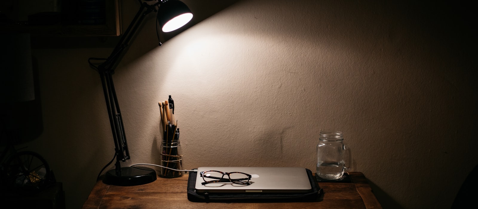 A desk lamp lighting a closed laptop, a pair of glasses and a jar of pens