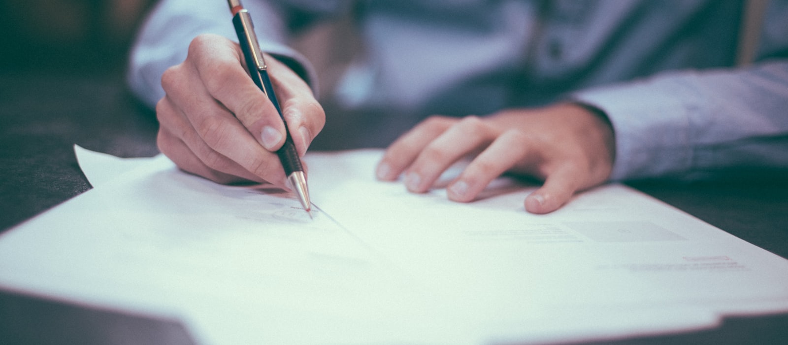 A person signing a typed document with a pen at a desk