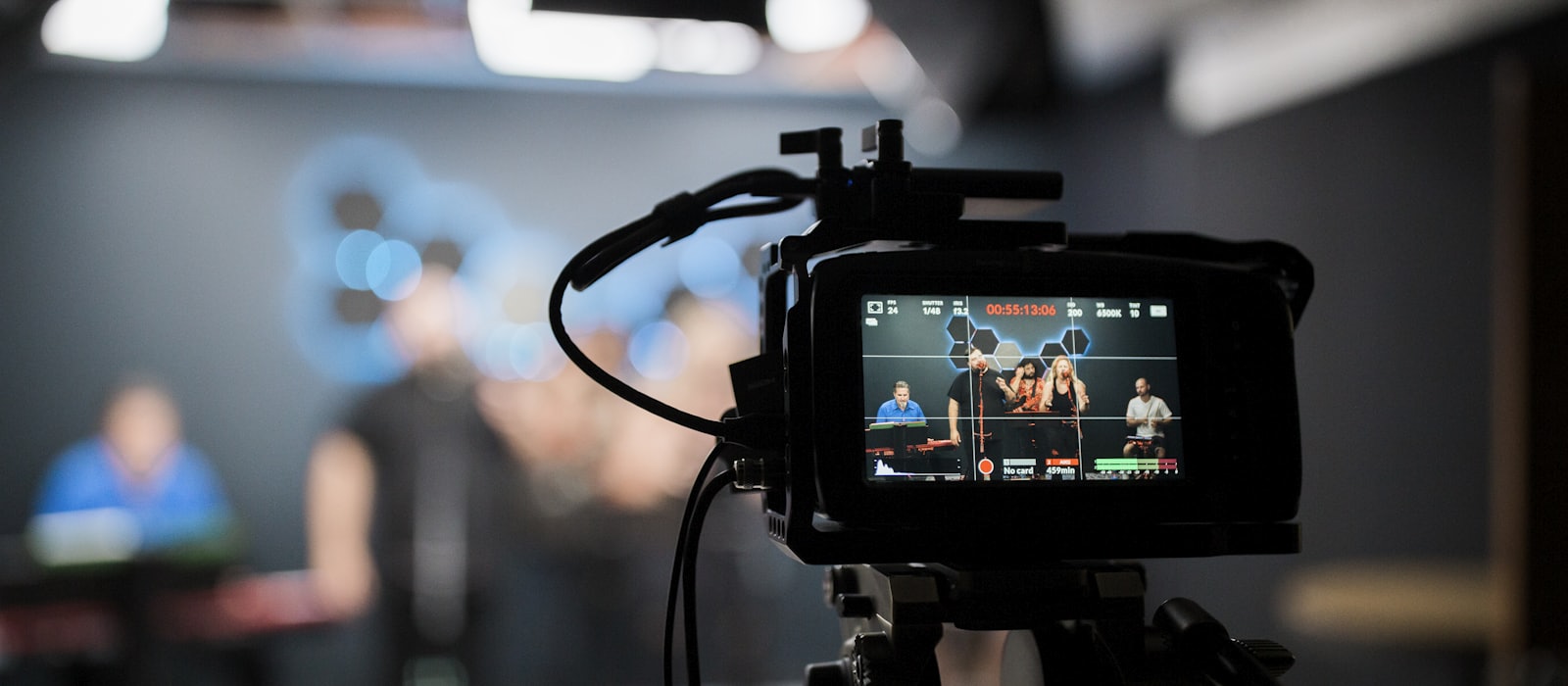 A broadcast camera on a tripod recording people in a studio