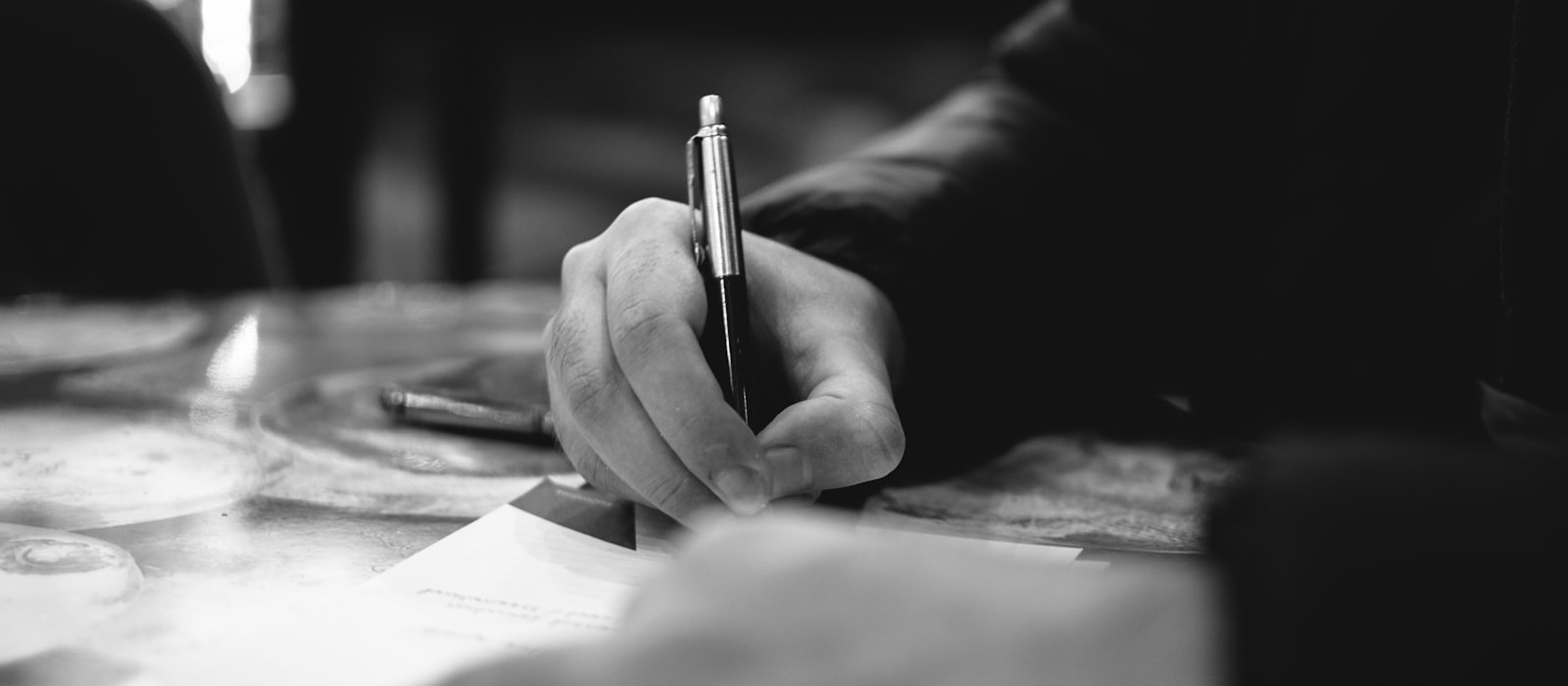 A hand writing on a document at a desk, in black and white