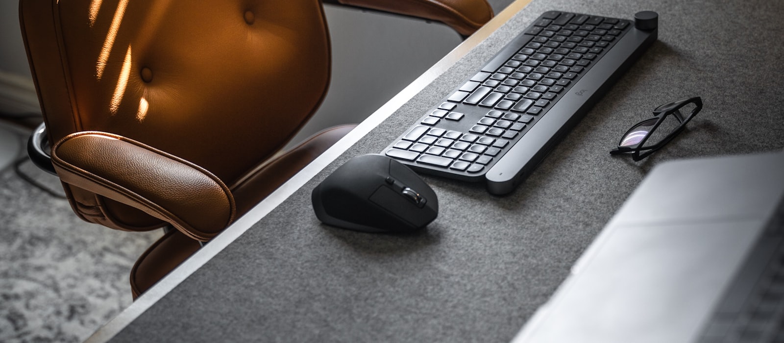 An empty leather office chair beside a desk with a keyboard, mouse and glasses