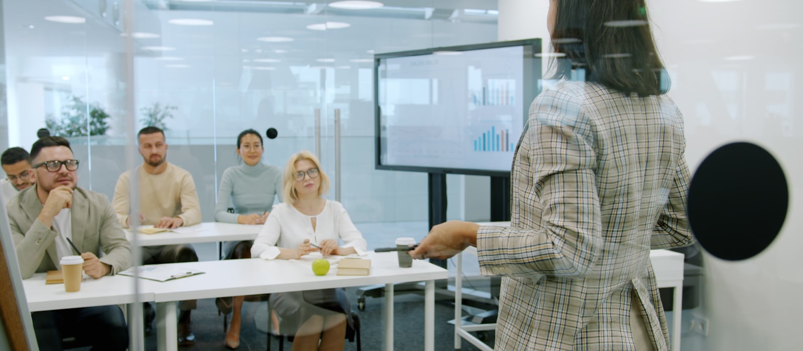 A person presenting to seated colleagues in a meeting room, beside a screen of charts