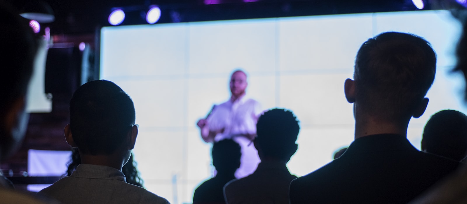 An audience seen from behind, watching a speaker on a large lit screen