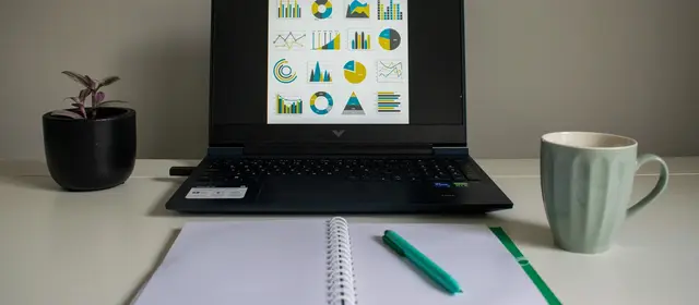 A laptop on a desk displaying a grid of charts, beside an open notebook and pen