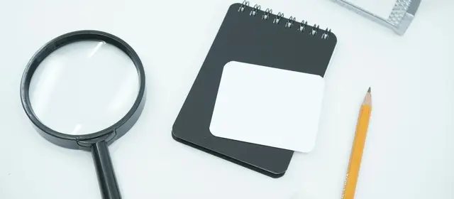 A magnifying glass, a small notebook and a pencil on a white desk
