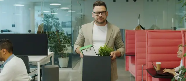 A man in glasses walks through an open-plan office carrying a box of his belongings while colleagues work at their desks