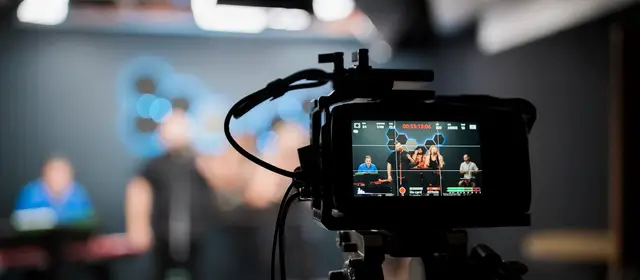 A broadcast camera on a tripod recording people in a studio