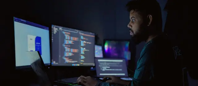 A software developer working at several monitors showing code in a dark room