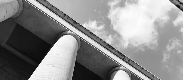Three stone columns under a cornice against a cloudy sky, in black and white