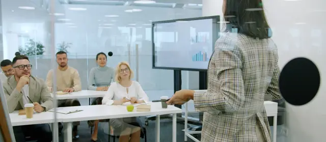 A person presenting to seated colleagues in a meeting room, beside a screen of charts