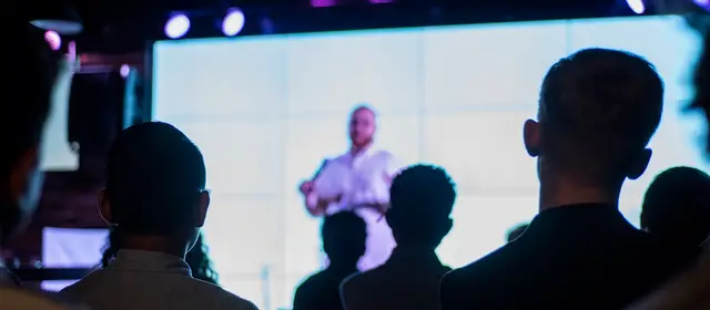 An audience seen from behind, watching a speaker on a large lit screen