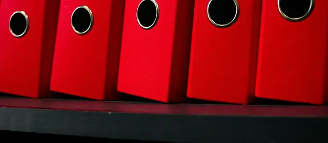 A row of red lever-arch binders standing on a dark shelf